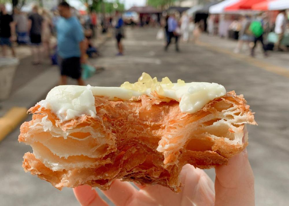 A hand holding a donut with white icing that is half eaten. Photo is taken outside at the farmers market in downtown fort wayne.