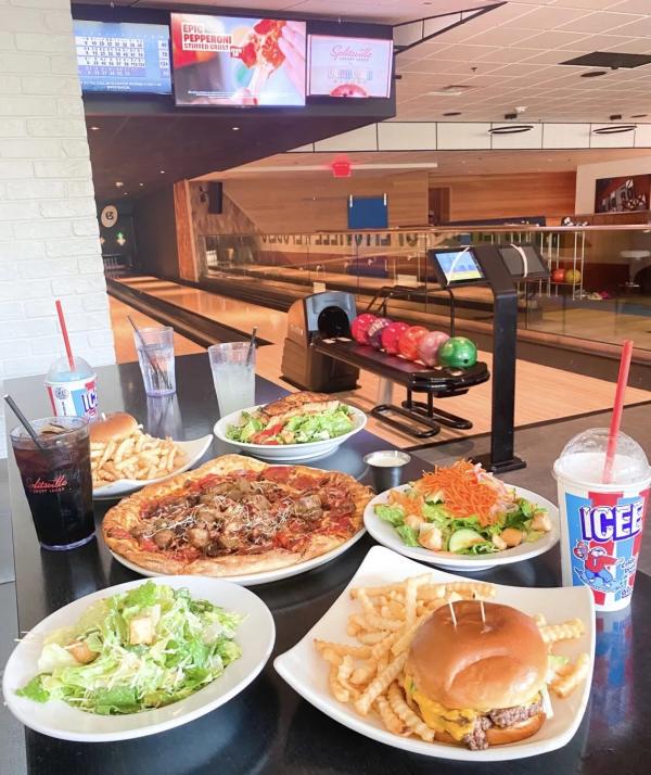 Image of a pizza, salad, hamburger, and two icees on a table. A bowling alley lane can be seen in the background.