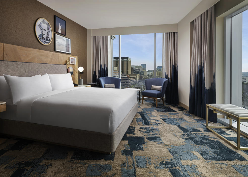 A room at This Otis Hotel featuring a large bed with white linens, blue carpeting and large windows.