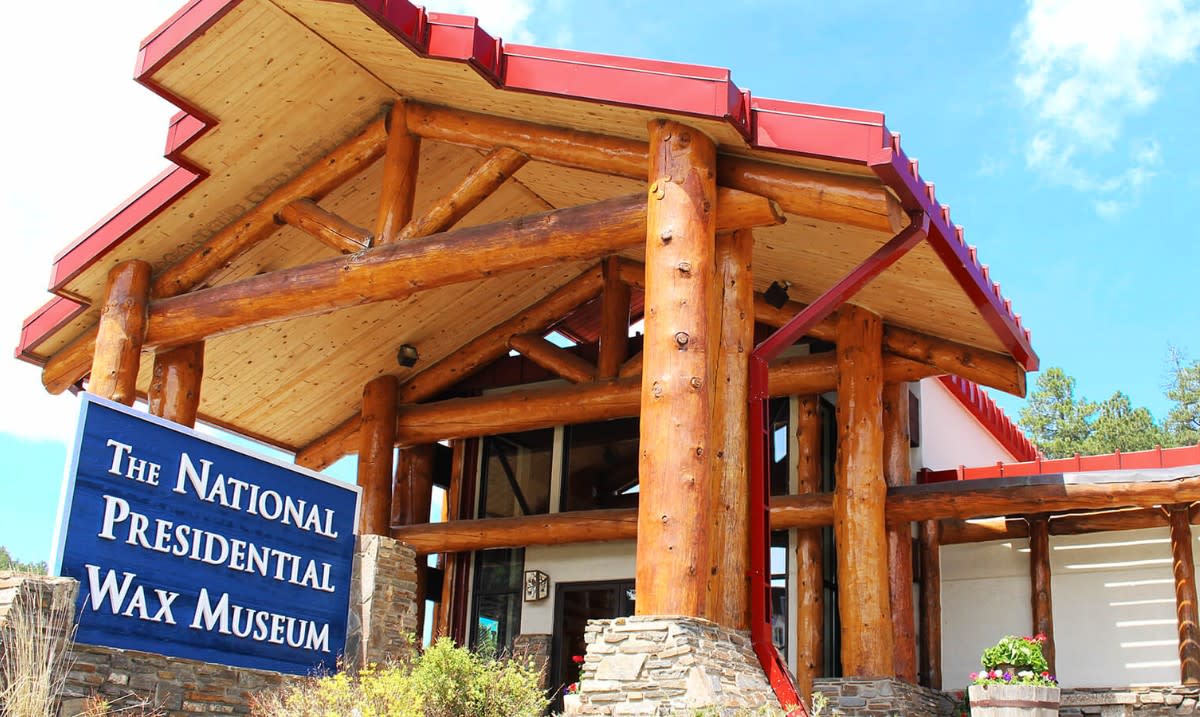 The exterior of the National Presidential Wax Museum noted by a blue sign with white text. It's a rustic building featuring stone supports and unfinished wood columns. The red roof is partially visible near the top of the photo.