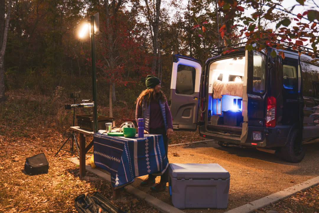 Person standing by a picnic table with camping gear and a lantern, next to an open van set up for sleeping in a wooded area at dusk.