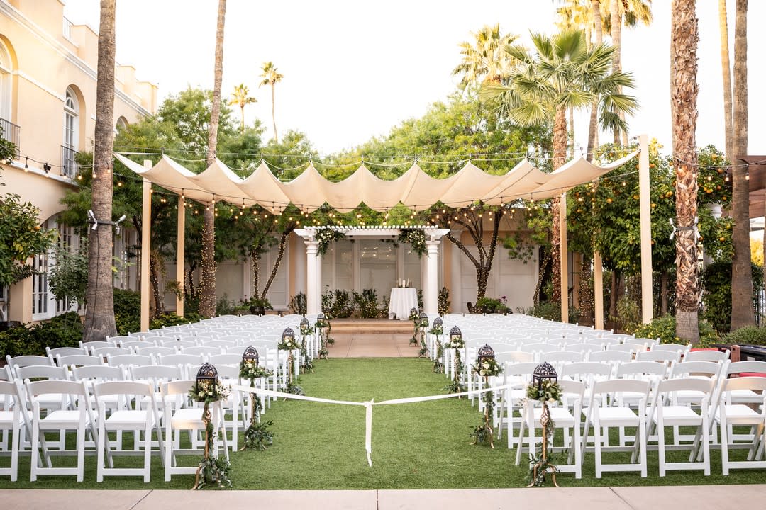 Palm tree & orange tree lined courtyard with several rows of white chairs sitting on grass, an isle down the center with white ribbon tied across, lantern style isle decor next to each isle side chair, a white ruffled canopy and lights above the chair, a white trellis as a gazebo with 2 stone stairs leading up, and a table with a white table cloth draped over it sitting under the gazebo.