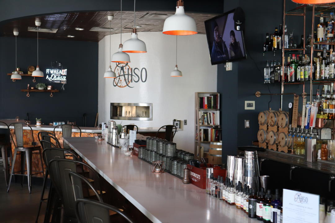 A large bar with black high-top chairs on one side and a wall of bottles and taps on the other at AhSo