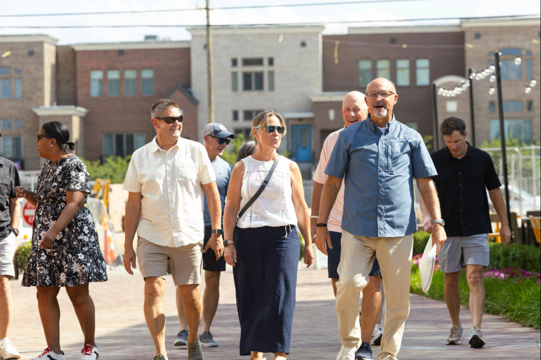 A group of people walks along a brick-paved street in downtown Bentonville, Arkansas, on a sunny day, with shops and landscaped areas in the background.