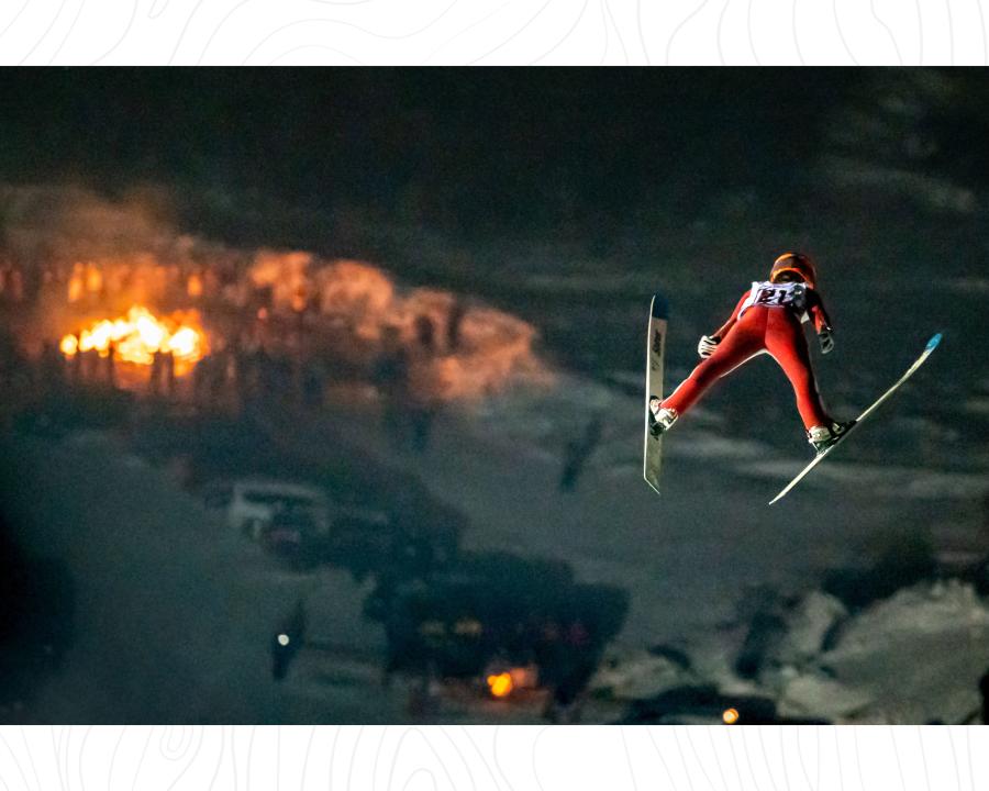 A skier suspended in air, looking over a crowd of spectators and giant bonfire at the Annual Ishpeming Ski Jumping Tournament in Ishpeming, MI