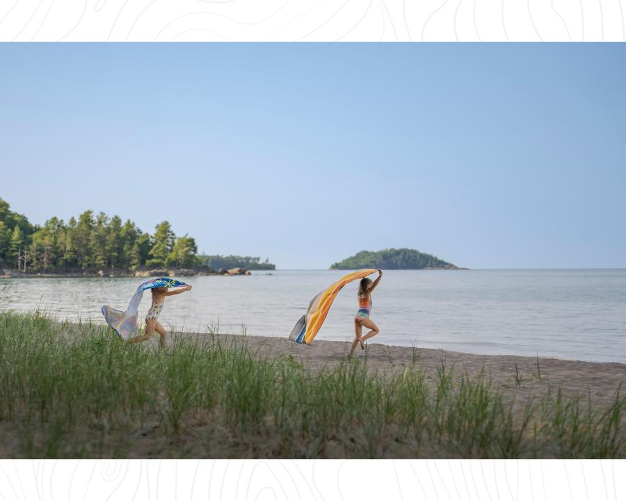 Two kids running on the sandy beach toward a calm Lake Superior on a summer day in Marquette, MI