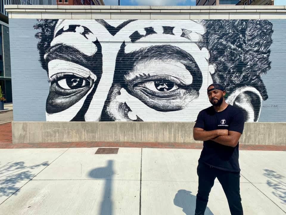 Artist Gee Horton standing in front of his mural of half a man's face painted in black and white