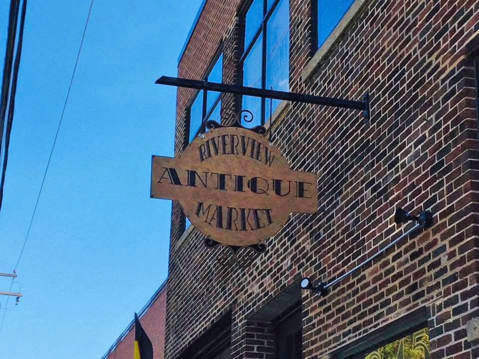 A hanging wooden sign for Riverview Antique Market extends from the brick exterior of the building. The vintage-style sign is mounted above the entrance, with large industrial windows and the historic brick façade visible against a blue sky.