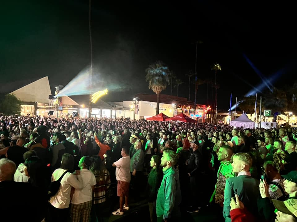 A large crowd of people gathered at night for an outdoor event, illuminated by stage lights with palm trees and tents visible in the background.