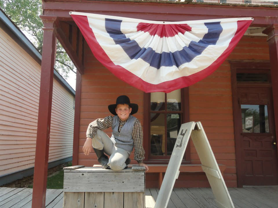 A young boy dressed in character sits in front of a building under a red, white and blue flag at Old Cowtown Museum