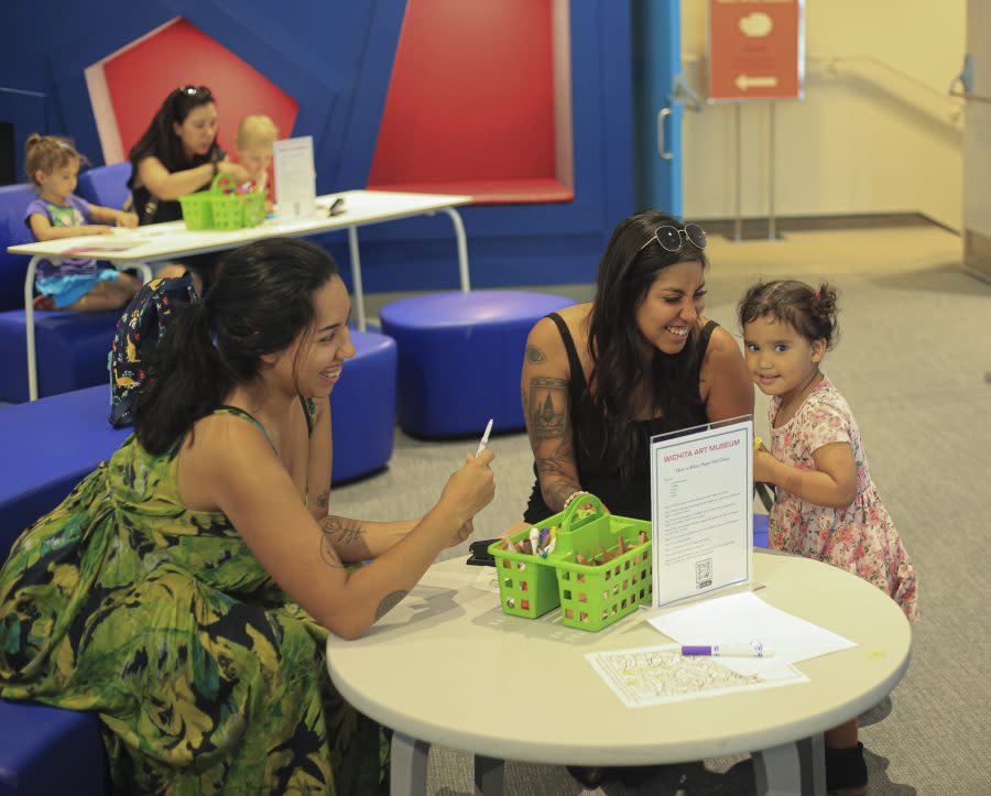 Two women smile at a girl creating art at a table during a Family ArtVenture event at Wichita Art Museum.