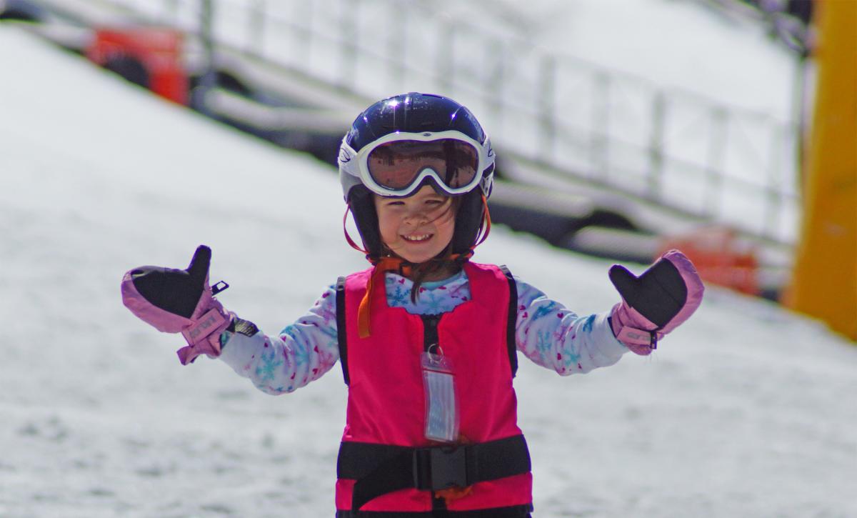 A young girl wearing ski gear smiles and holds up her glove-covered hands 