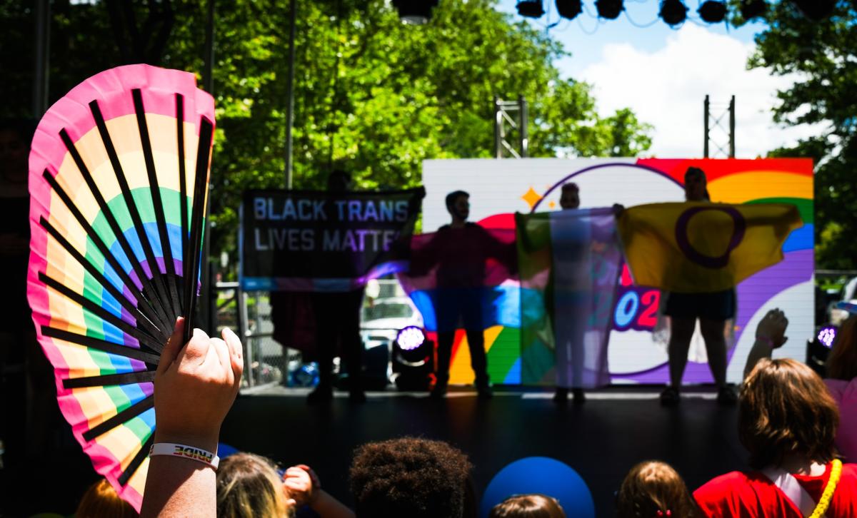 Photo taken at the Lawrence PRIDE street party, featuring a hand holding a rainbow fan in the foreground and performers on stage holding pride flags in the background.