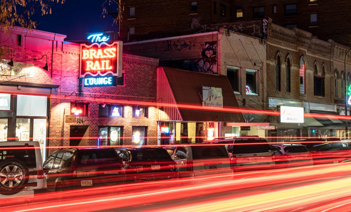 Nighttime street view with light trails from passing cars. A neon sign reads "The Brass Rail Lounge."