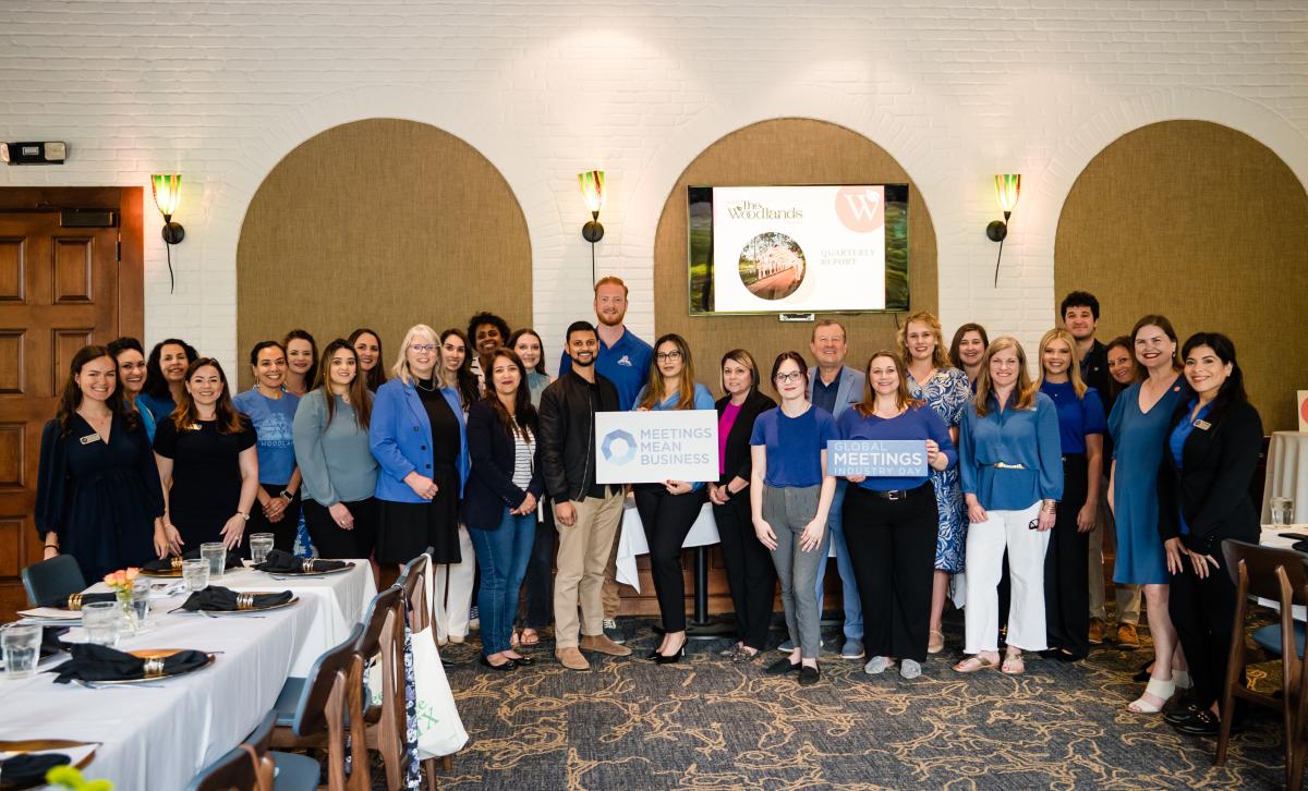 Twenty-eight people smile at the camera for a group photo. There are men and women of all ages, all dressed business-casual, and most dressed in royal blue in honor of GMID. The photo is taken indoors, in the Black Walnut private dining room. Two women in front hold signs that read, "MEETINGS MEAN BUSINESS" and "GLOBAL MEETINGS INDUSTRY DAY." On the wall behind the group is a TV picturing the title slide of VTW's 2024 Q4 Report.