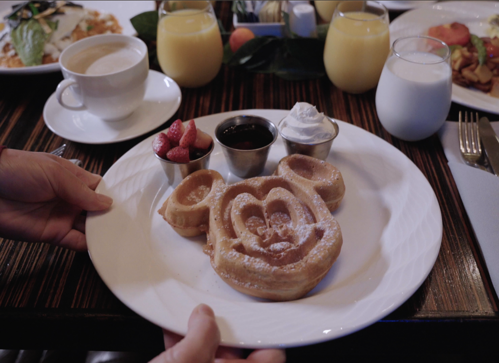 Image of a plate of waffles shaped like Mickey Mouse. Plate is placed on a dark brown/black wood table top. There is also a cup of coffee in the top left-hand corner, and a glass of milk and orange juice in the top right-hand corner. There are also two additional plates of food place in both top corners.