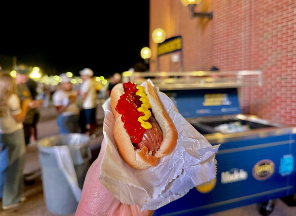 Person holds a hot dog with katchup and mustard zig-zagged on top, outside of a brick stadium.