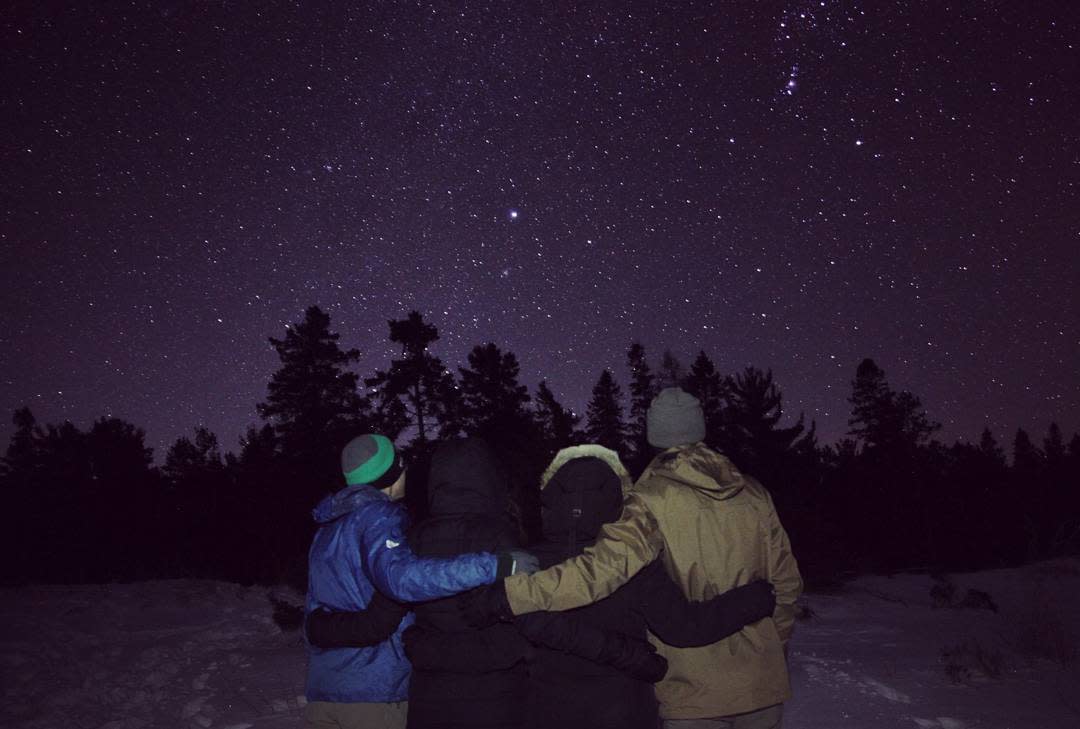 Four people stand arm and arm in the snow and gaze up at a starry night sky above Mackinac Island