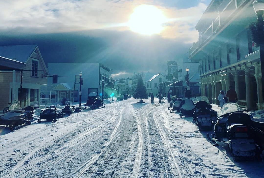 Sun shines on snowy Mackinac Island Main Street with snowmobiles parked along both sides and tracks down the middle