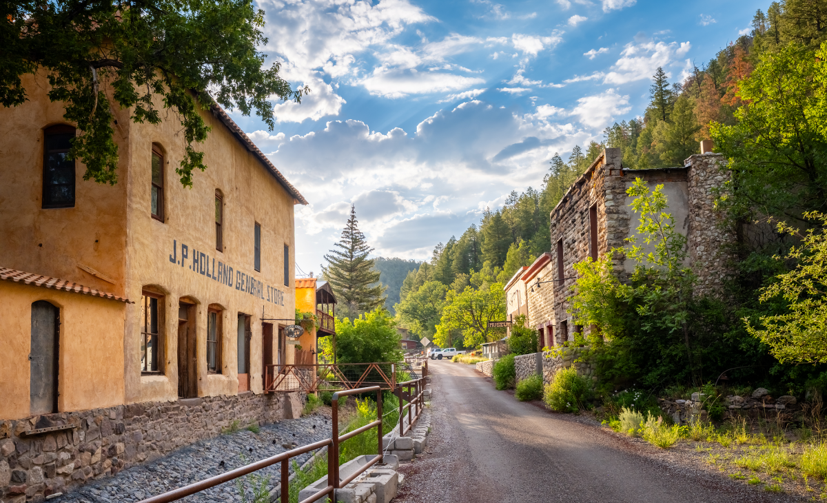 Historic buildings line the narrow main street of Mogollón, New Mexico, under a bright blue sky surrounded by forested canyon walls.