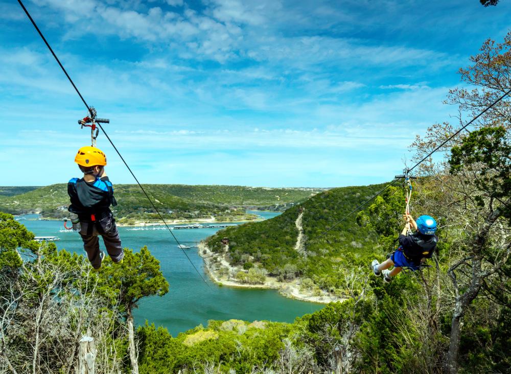 Two children flying down parallel zip lines towards rolling hills and Lake Travis.