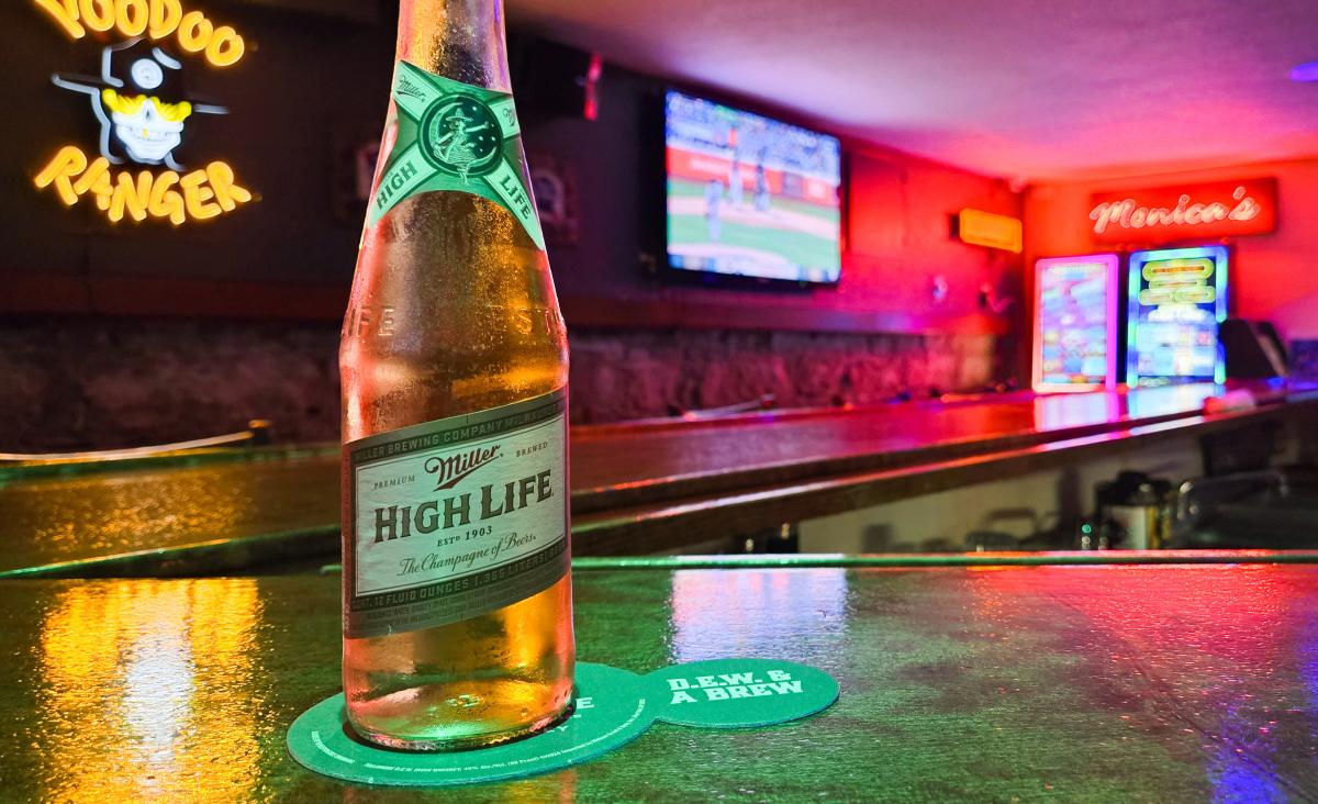 A bottle of Miller High Life beer sits on a green bar counter at Monica’s on Astor in Milwaukee. Neon signs, including a Voodoo Ranger logo and a glowing red “Monica’s” sign, illuminate the background. A TV on the wall shows a baseball game, adding to the relaxed bar atmosphere.