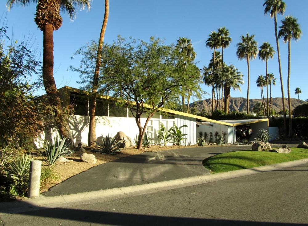 A mid-century modern house with a flat roof and clean lines surrounded by palm trees and desert landscaping under a clear blue sky with mountains in the background.