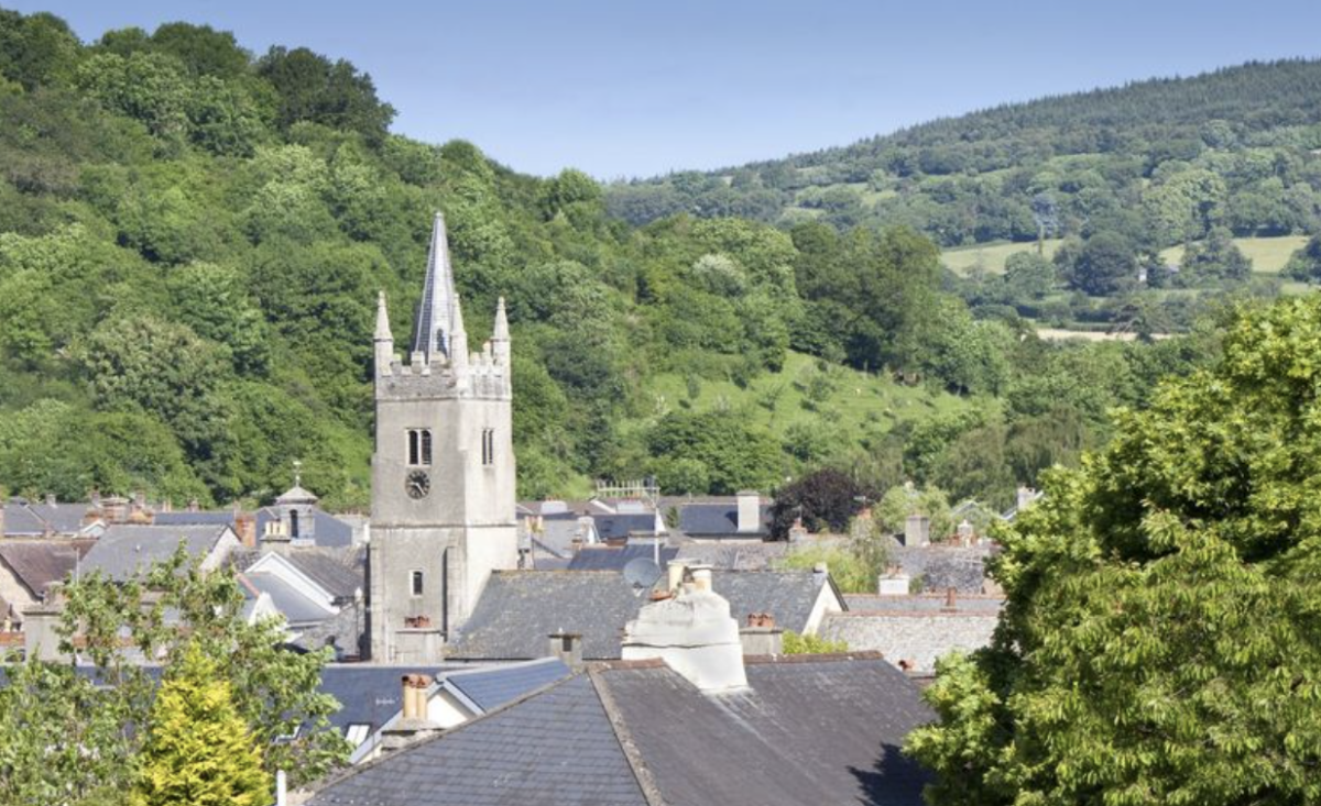 A scenic view of a quaint village, dominated by a stone church tower with a spire, surrounded by lush green hills under a clear blue sky.
