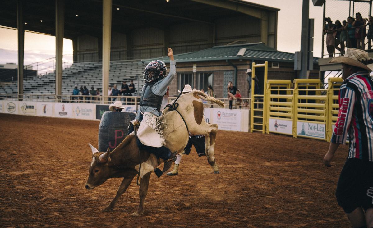 Rider on a bucking bull at a rodeo, with spectators and barrier fences visible in the background at dusk.