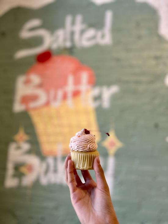 A hand holding a cupcake in front of an artistic sign that says "Salted Butter Bakery"