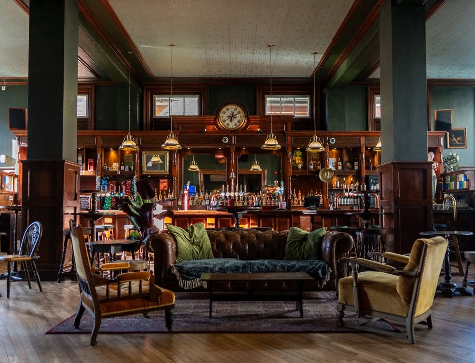 Interior of The Queen Mary pub with leather coach, stylish yellow armchairs and bar area with hanging pendant lights