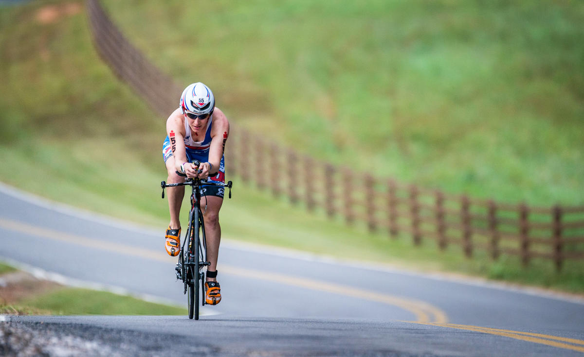 Chattanooga Ironman participants rides bike on road with field behind them.
