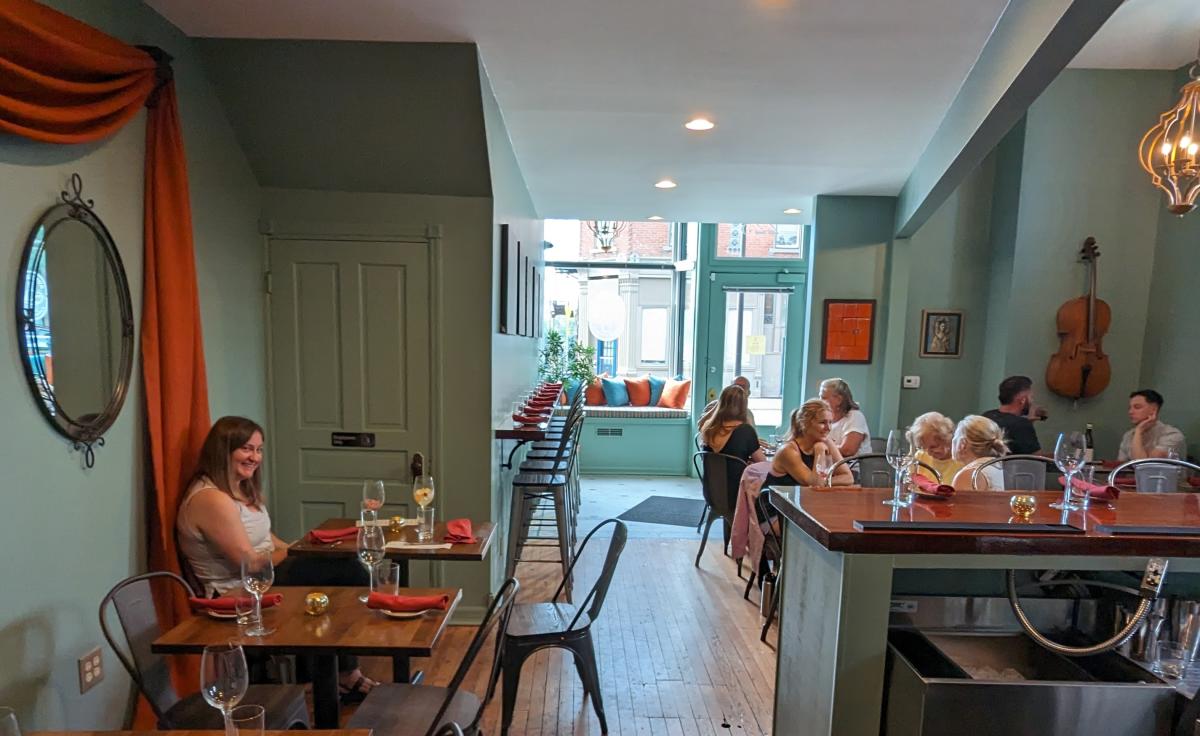 Image is of inside the restaurant with tables and chairs to the left, bar to the right and people seated at tables in the background.