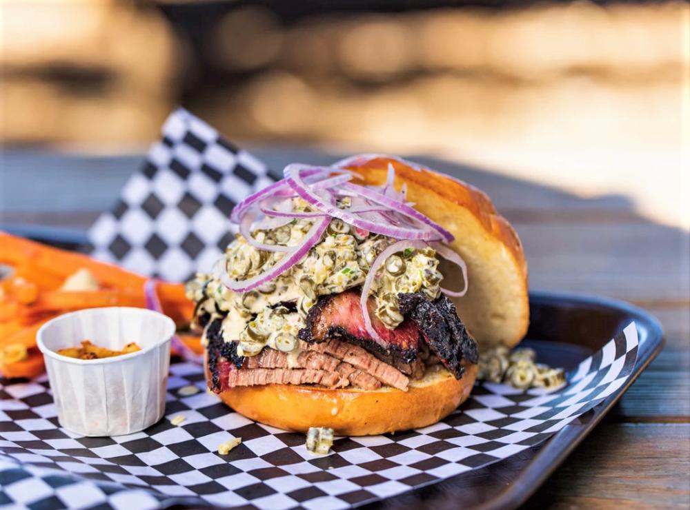 Sliced brisket sandwich piled high with a vegetable slaw, thinly sliced red onions, and topped with a toasted, buttered bun.