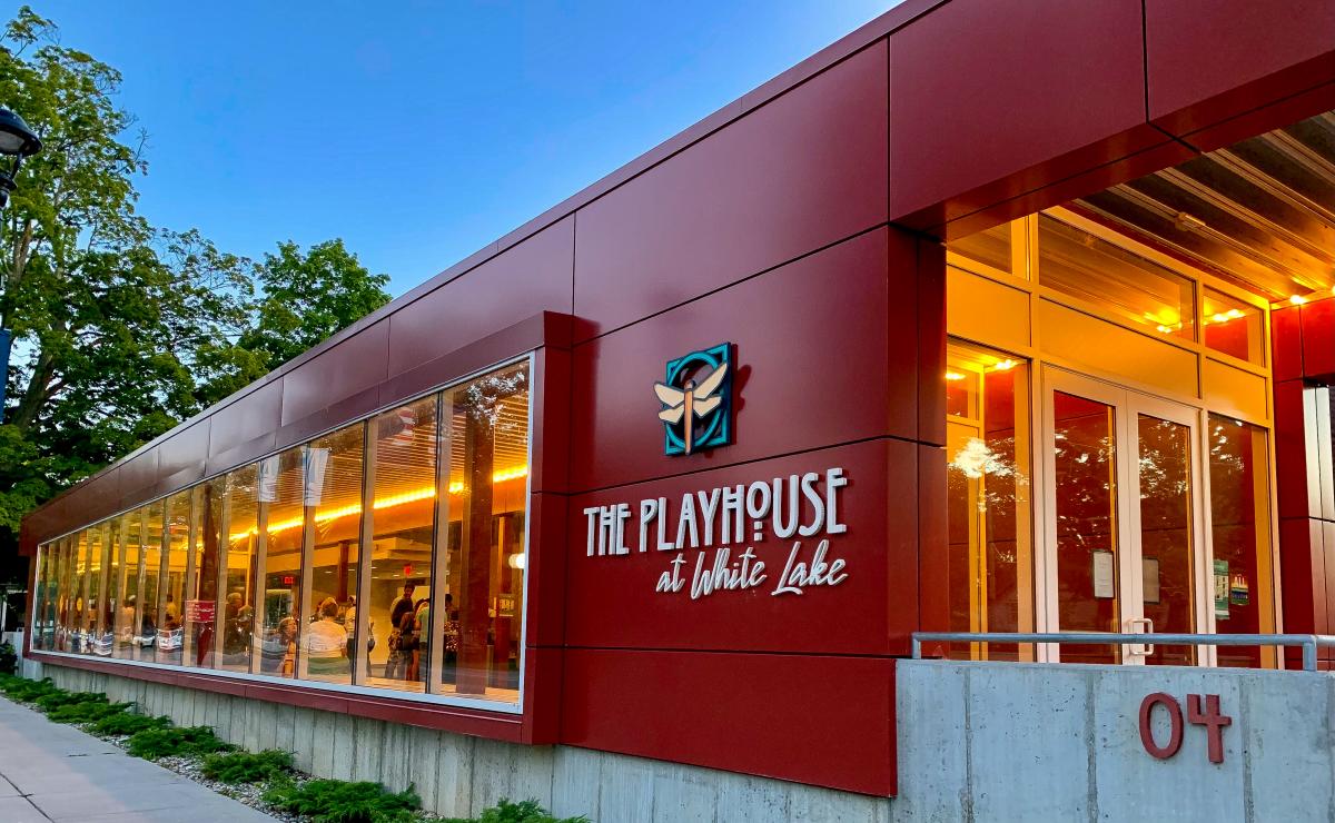 dark red building with warm windows running along side and at entrance. Sign says the playhouse at white lake with dragonfly logo