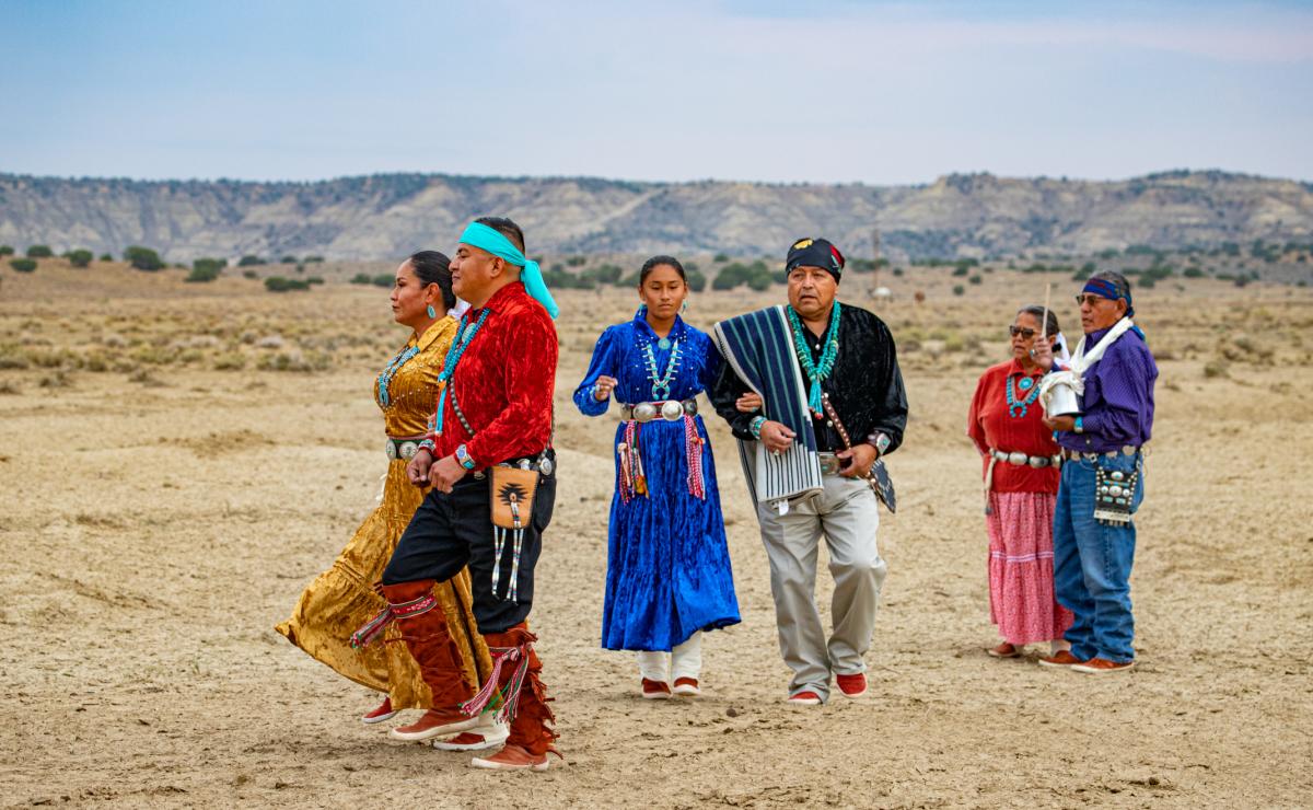 Group of Native American individuals in traditional attire participating in a cultural ceremony in a desert landscape.