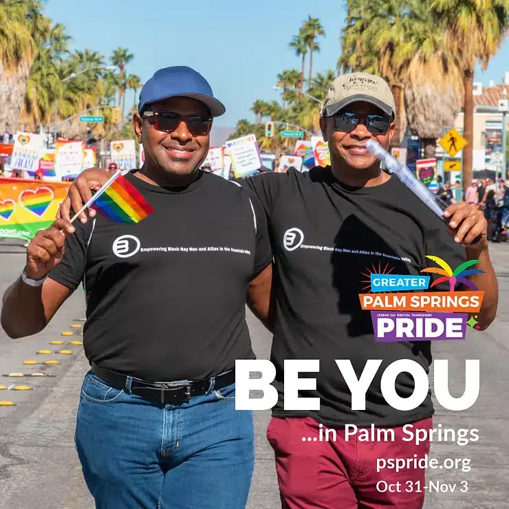Two smiling men walking arm-in-arm at a pride event, one holding a rainbow flag, with palm trees in the background and text promoting Greater Palm Springs Pride event dates.