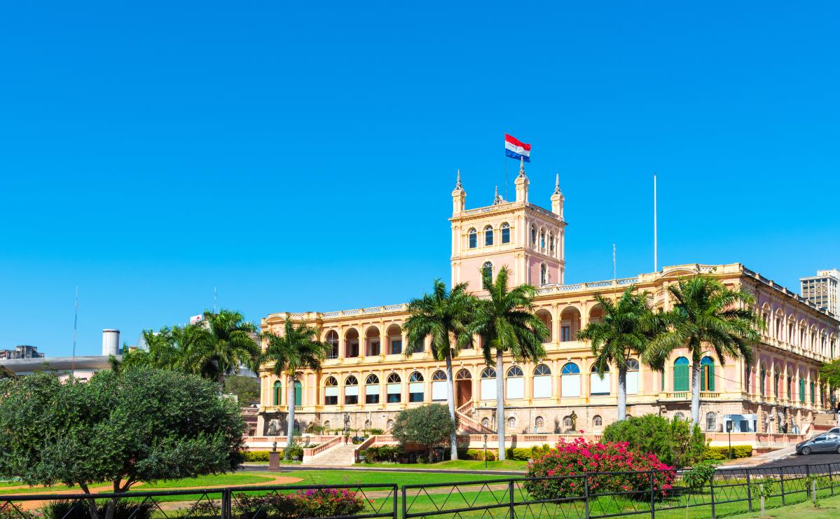 A large, ornate building with a central tower and a Paraguayan flag flying from its top. The building is surrounded by palm trees and a manicured lawn.