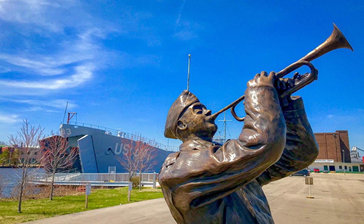 Bronze statue of ww2 soldier playing trumpet with ww2 landing ship docked in distance