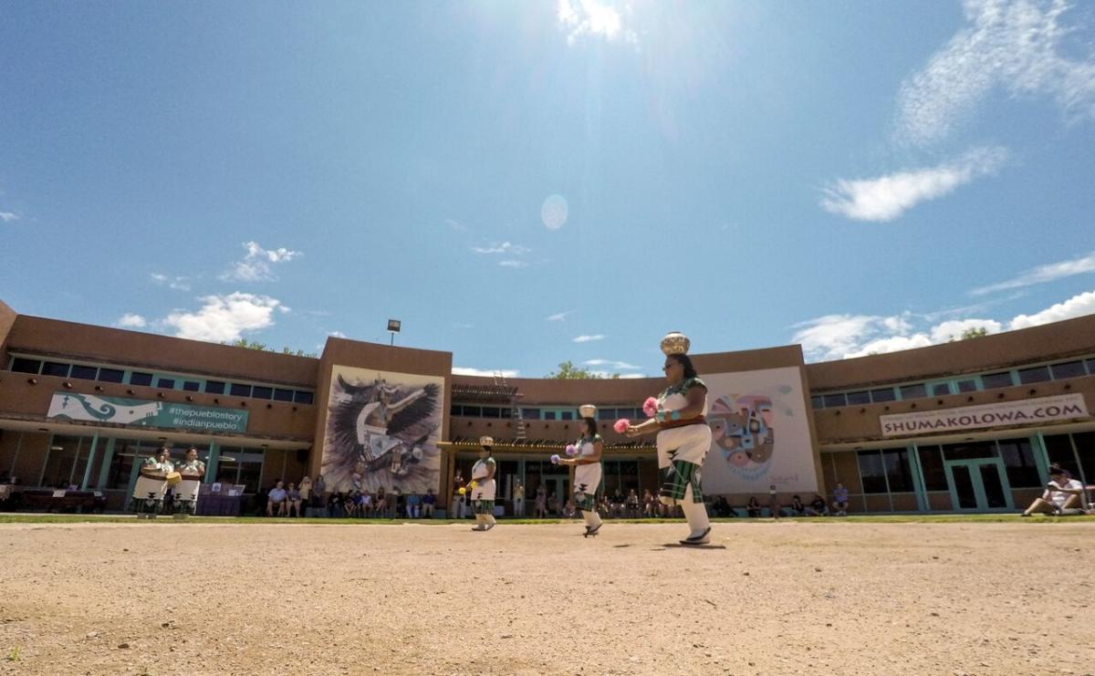 three people dressed in traditional indigenous attire perform a dance in the middle of a dirt pavilion