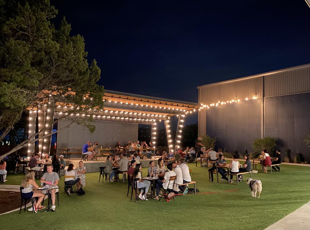 outdoor lawn at Meanwhile Brewing Co at night, with a stage and twinkle lights behind tables with people