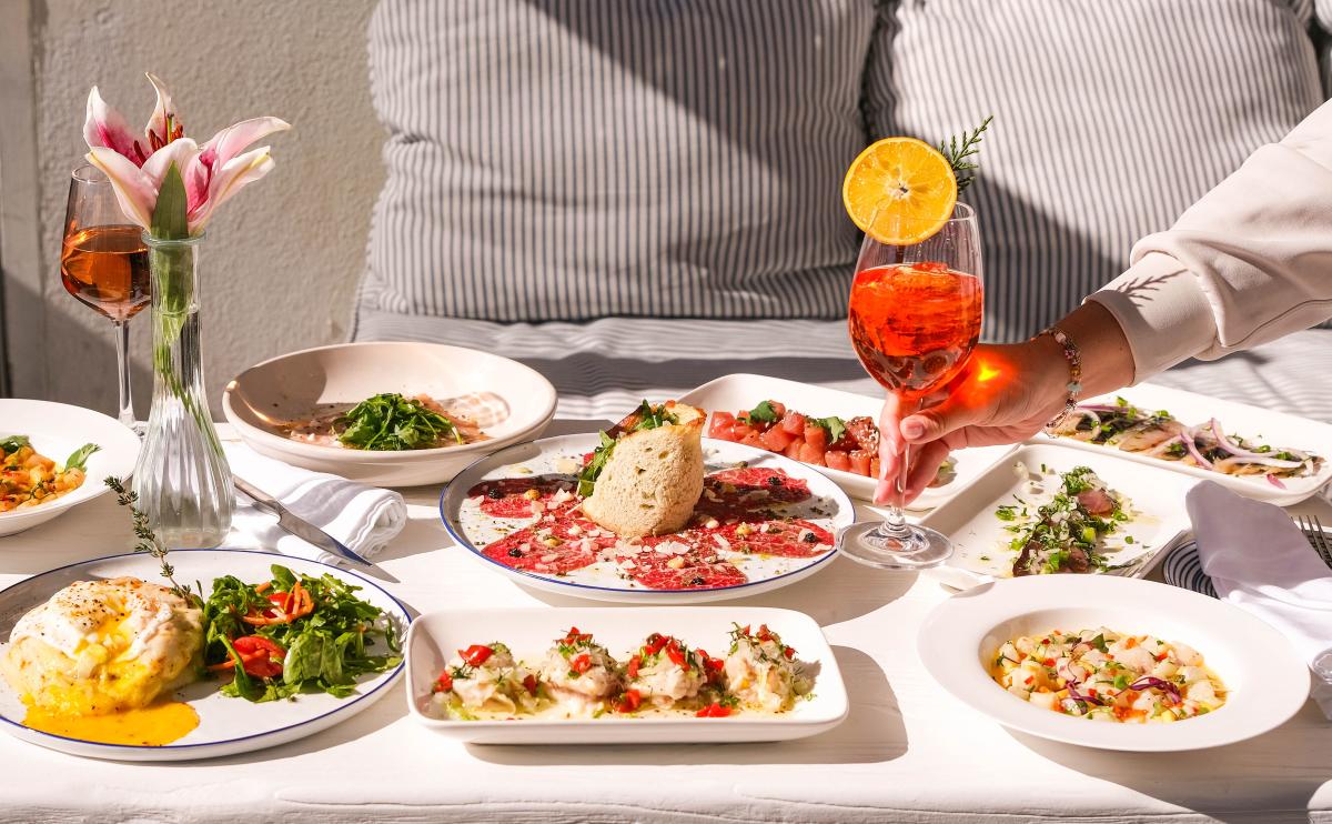 Image of a table at Yamas with plates of food, flowers and a hand reaching across the table picking up an Aperol Spritz drink.