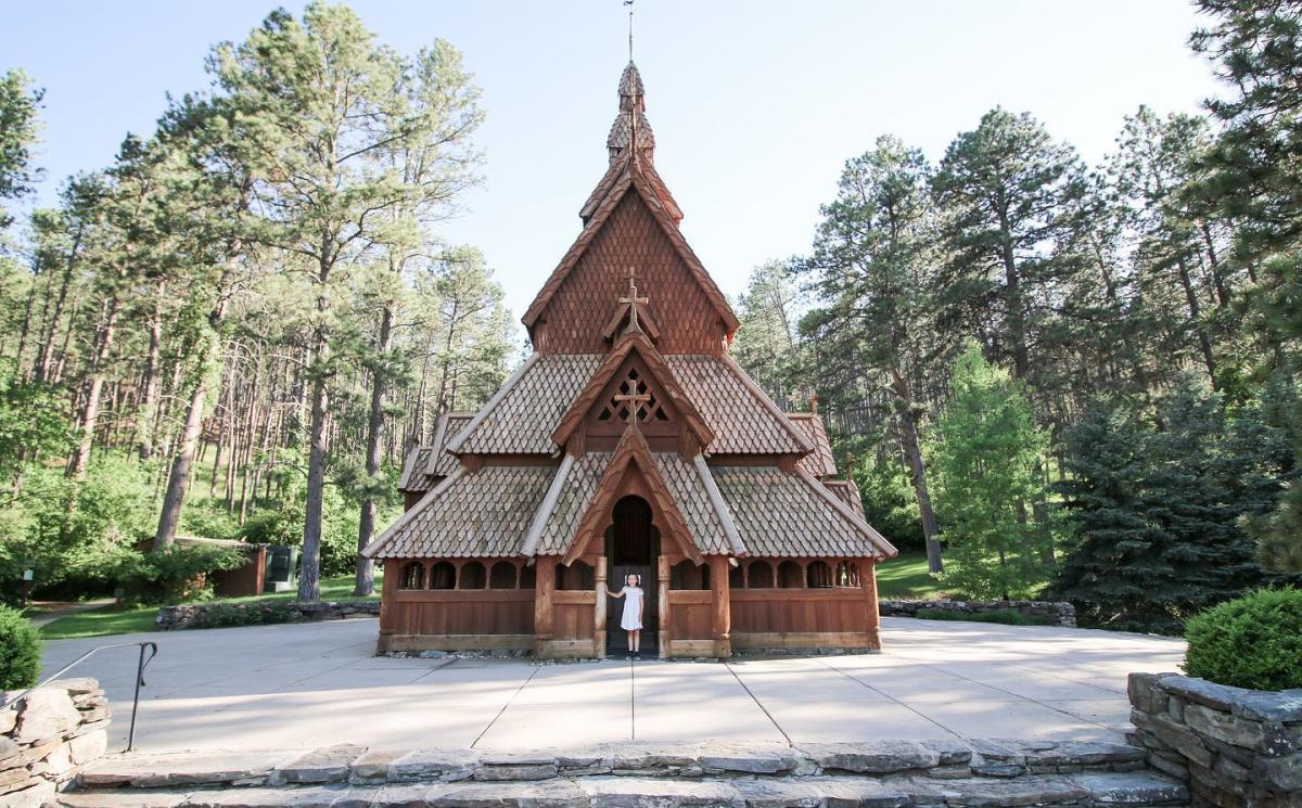 A wooden stave church with intricate carvings stands amidst a lush forest. A person in white stands at the entrance, conveying a sense of tranquility.