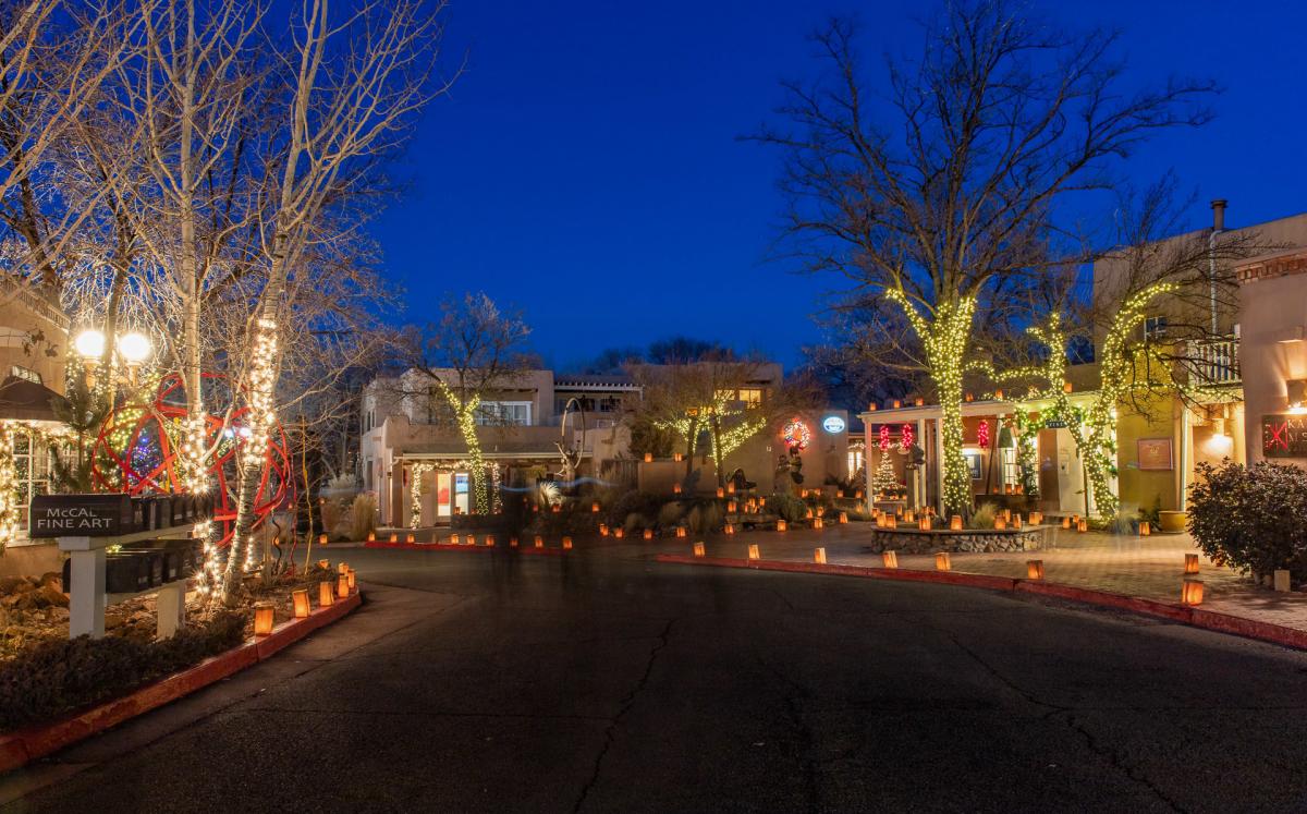 A beautifully lit street scene at dusk in Santa Fe, showcasing decorative lights wrapped around trees and buildings. Warmly glowing lanterns line the pathways, creating a cozy and festive atmosphere in a district known for its art galleries.
