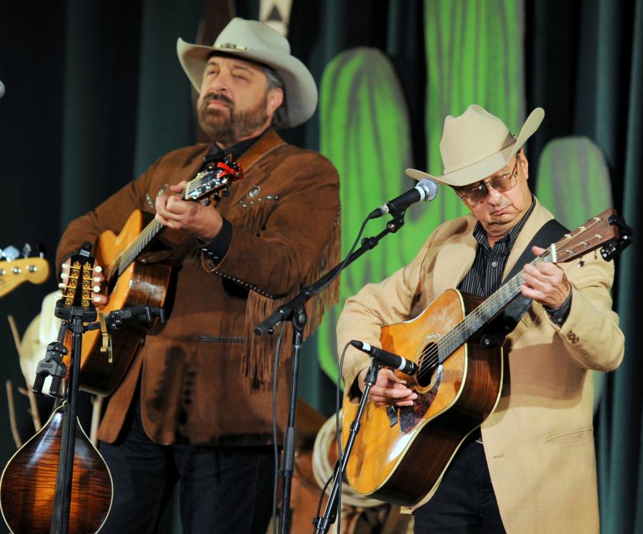 Western musicians and cowboys Jon Chandler and Ernie Martinez performing with guitars on stage.
