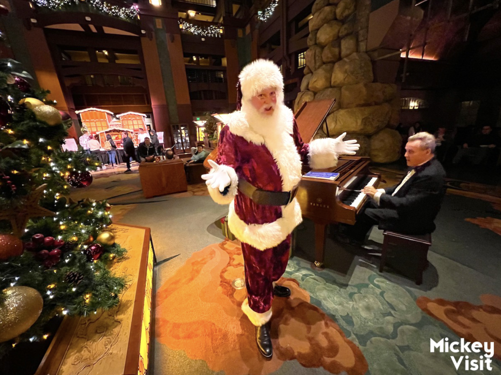 Image of Santa Claus dressed up in his red and white holiday attire. He can be seen posing for the camera, standing next to a piano player. To his left is a Christmas tree decorated with red, silver, and gold ornaments.