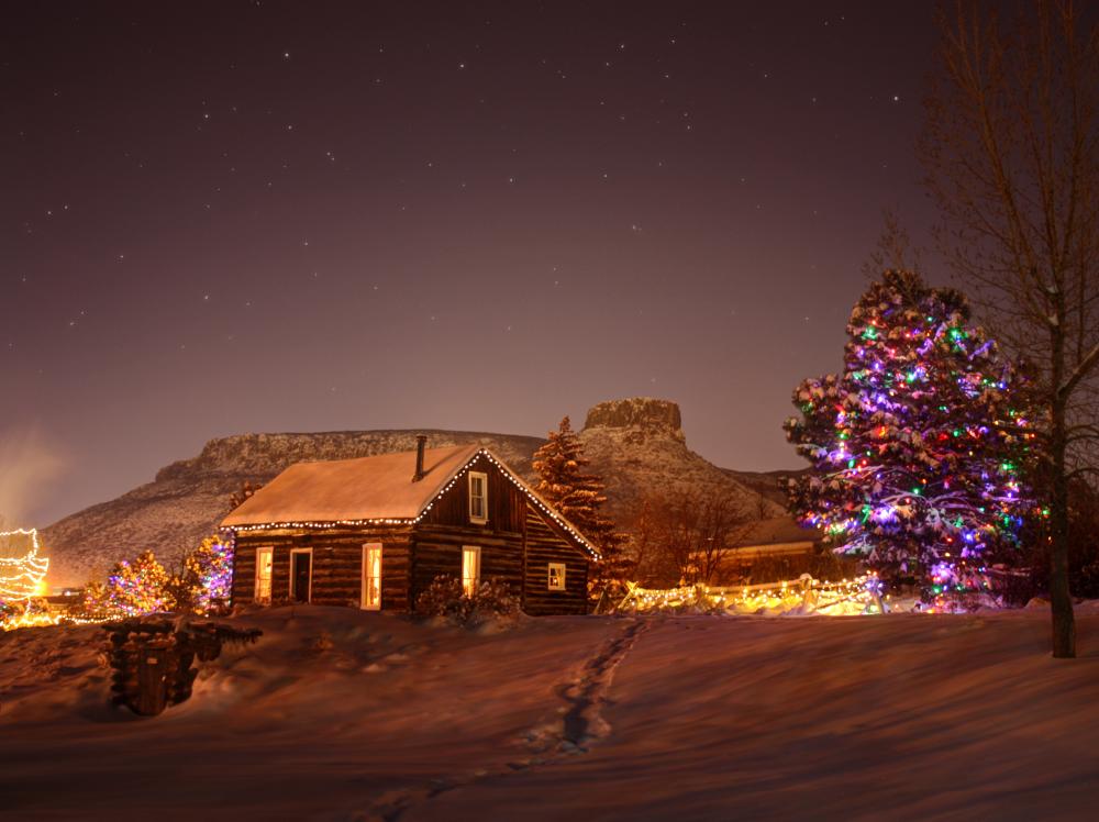 A snow covered Golden History Park at night with lights on inside the log cabin and trees in the park, castle rock is visible in the background