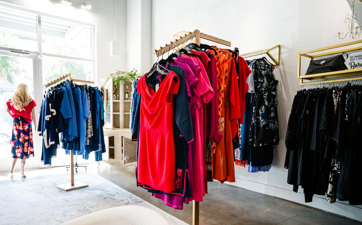 The camera overlooks the interior of Bowtique. It features multiple racks of classy, colorful clothes for various occasions (formal dresses, sundresses, sleepwear, etc.). Standing at the front door with her back to the camera is a mannequin modeling a bright pink blouse and a skirt that is navy with bright pink flowers.