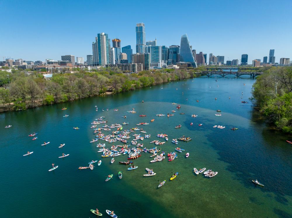 Colorful kayaks and paddleboards on blue-green water with a view of the Austin skyline in the background.