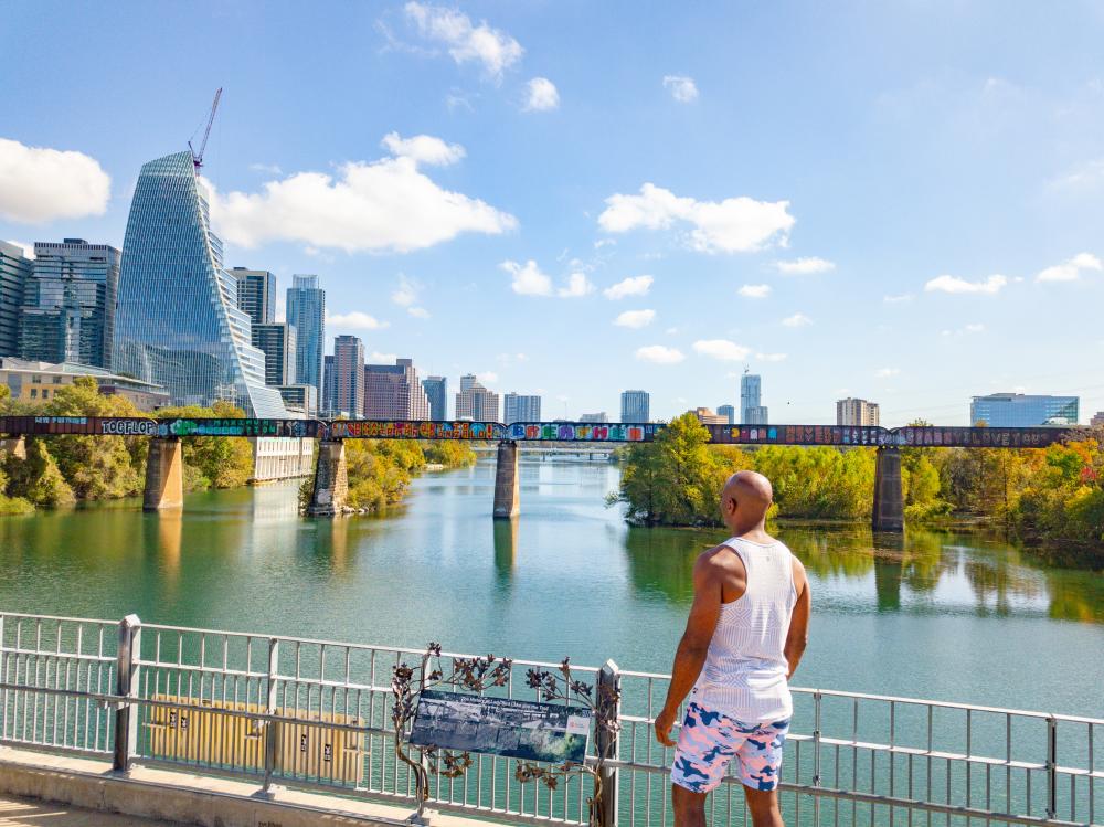 Man walking on Pfluger Pedestrian Bridge on the Hike & Bike Trail, looking across the water at the downtown skyline.